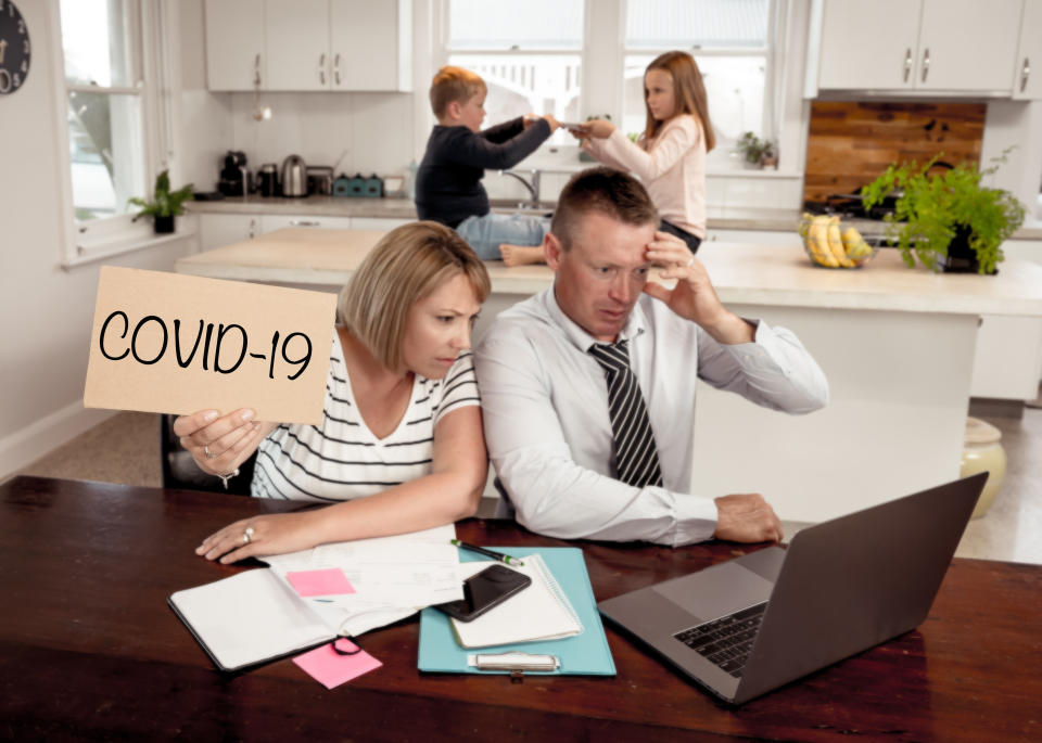 Mother holds up 'COVID-19' sign while concerned father looks at a laptop and children fight in the background.