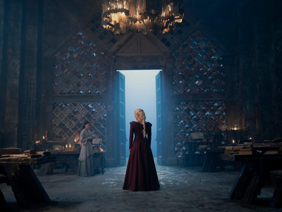 emma d'arcy as rhaenyra targaryen on house of the dragon, seen from the back wearing a red dress and framed in a shot of a large, open door. to rhaenyra's left is sonoya mizuno as mysaria, wearing blue robes and standing near a table