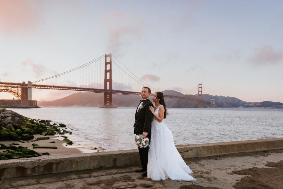 A bride touches her groom's shoulder as they pose in front of the Golden Gate bridge.