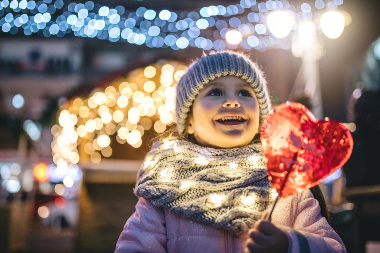 A child in winter clothes holds a heart-shaped lollipop surrounded by holiday lights. Donating to the Give a Christmas program can help children from needy families enjoy the holidays more.