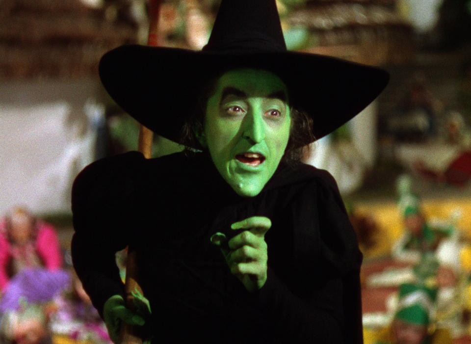 Margaret Hamilton dressed as the Wicked Witch of the West, with green face makeup and wearing a black outfit and pointed hat, from "The Wizard of Oz"