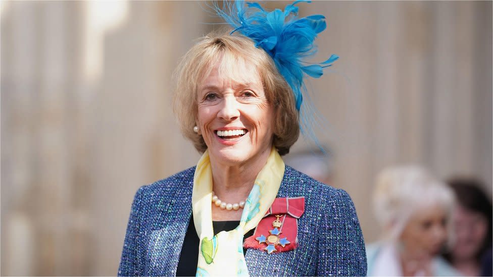 Dame Esther smiles, dressed up with a feathered blue fascinator