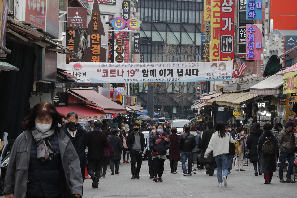 People wearing face masks walk through a market in Seoul, South Korea, Thursday, March 4, 2021. South Korea's central bank says the country's economy shrank for the first time in 22 years in 2020 as the coronavirus pandemic destroyed service industry jobs and depressed consumer spending. The banner reads: "Lets' overcome Corona 19 together." (AP Photo/Ahn Young-joon)