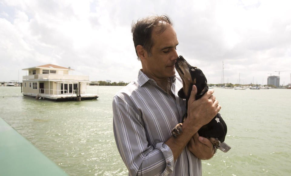 FILE- In this April 22, 2014 file photo, Fane Lozman poses for photos holding his dog near his home floating in the waters near North Bay Village, Fla. Four years ago, Fane Lozman caught lightning in a bottle when the U.S. Supreme Court agreed with him that his floating home was a house, not a vessel subject to seizure by a Florida city. However, since that January 2013 decision, Lozman's legal battle with Riviera Beach has continued because of the city's refusal to reimburse him for $200,000 in legal fees and the estimated $165,000 value of the floating home the city had destroyed. (AP Photo/J Pat Carter, File)