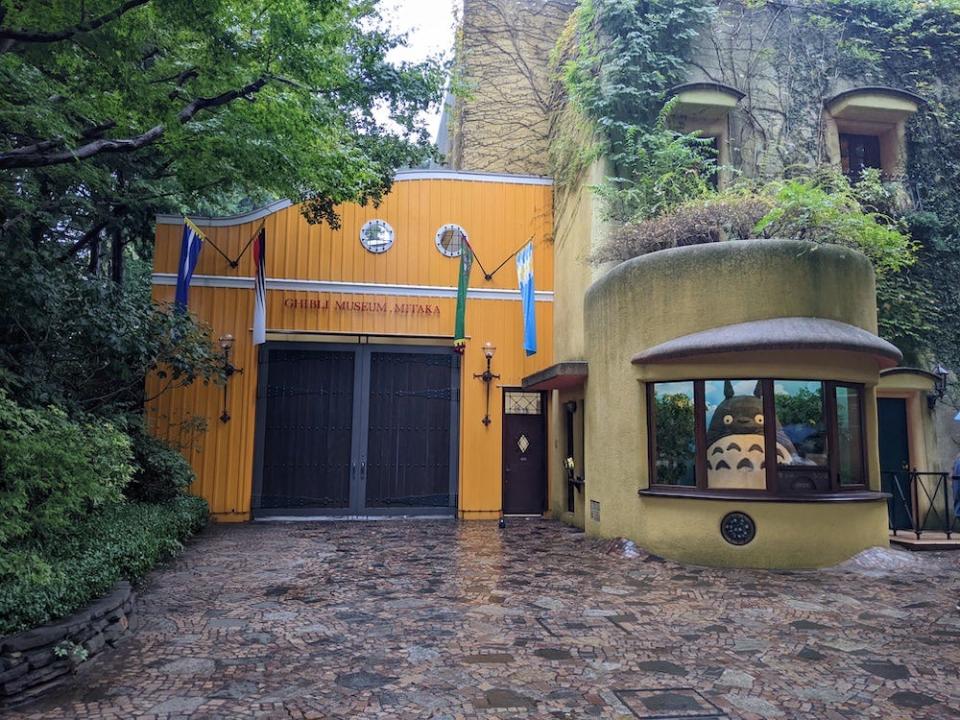 Image of the front of the Ghibli Museum. The left side is painted yellow with colorful flags hanging in a row. On the right is a circular building with a glass storefront and a giant plush Totoro figure.