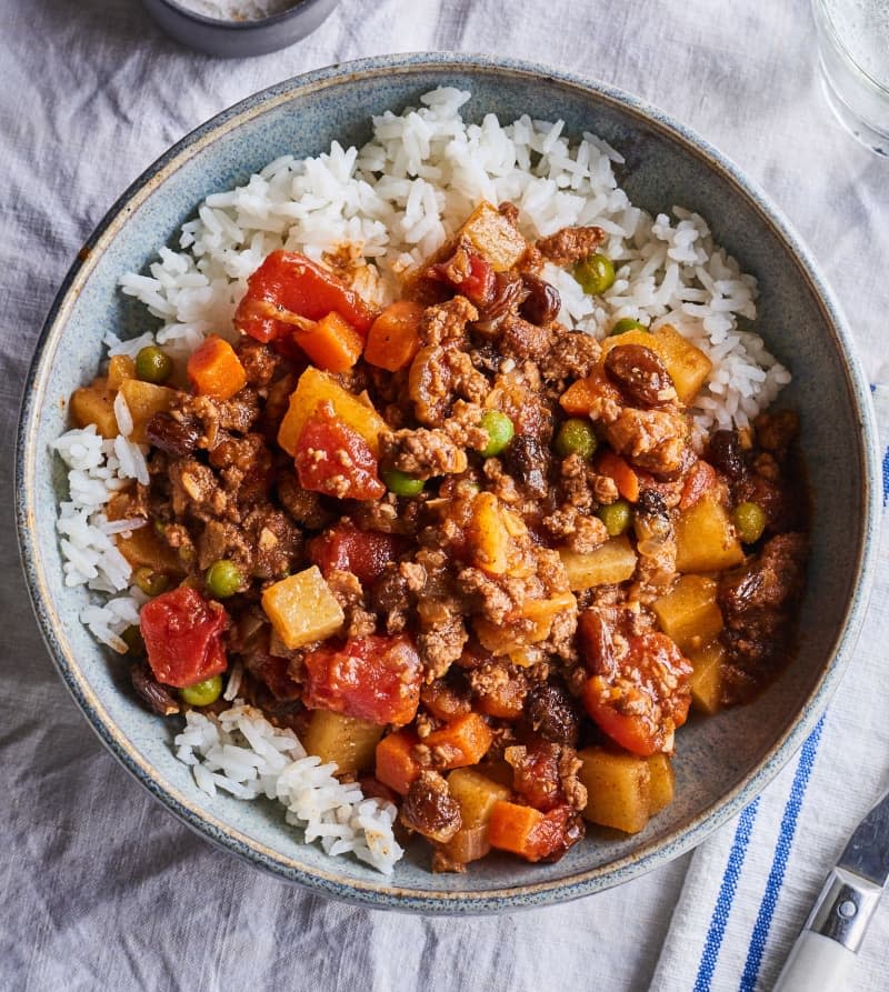 picadillo sits in a bowl over rice next to a glass and a fork