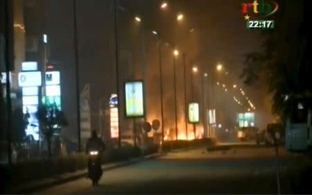 A motorcyclist rides along a road while fire and smoke rising from Splendid Hotel in Ouagadougou where suspected Islamist fighters are holding hostages in this still image from a video grab, January 15, 2016. REUTERS/RTB via REUTERS TV