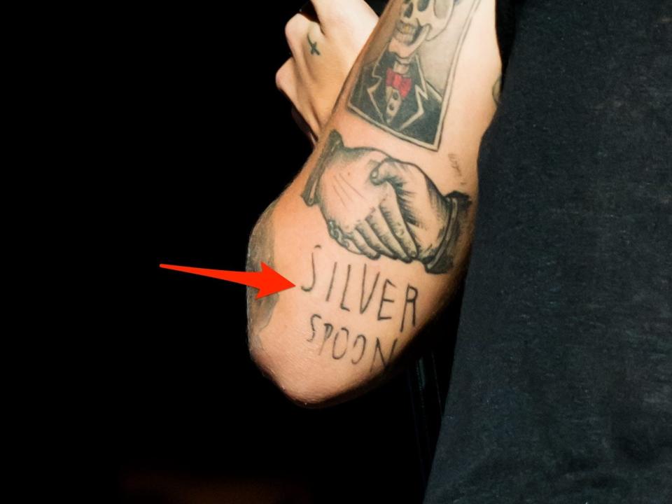 A red arrow pointing to Harry Styles' "silver spoon" tattoo on the back of his left arm.