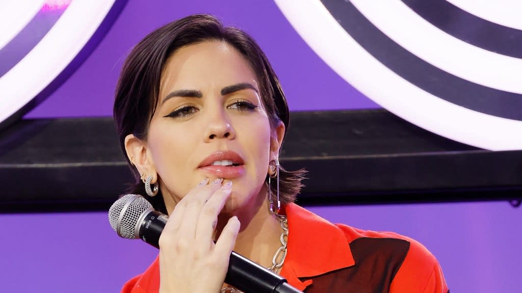 Katie Maloney in a red top, biting her lip and holding a microphone