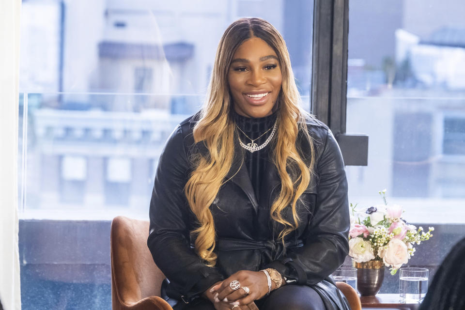 Serena Williams in a black jacket sits in a chair in front of a window in the city.