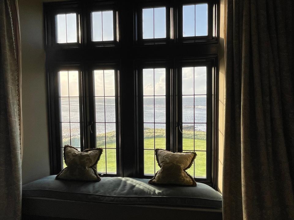 Windows overlooking sea and grass with a bench and pillows in foreground
