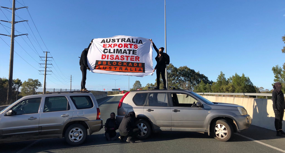 Protesters in black stand on top of two cars that are blocking a road. It reads: Australia exports climate disaster.