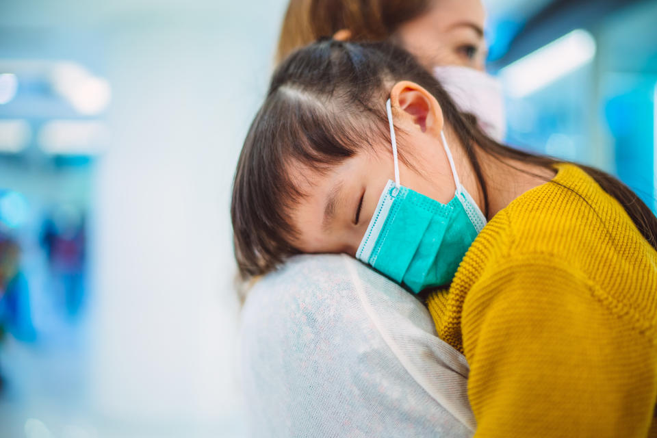 Little daughter with medical face mask sleeping on mom's shoulder in shopping mall. The young mom is taking care her sick daughter while strolling in shopping mall.