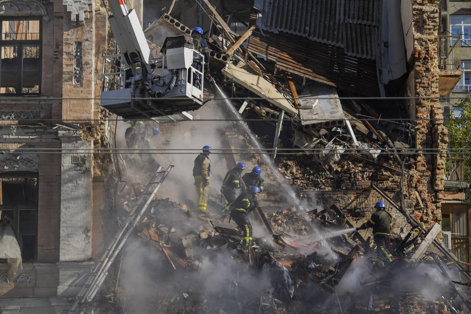 Under a crane, rescuers in hard hats climb down a huge pile of steaming rubble in a destroyed residential building, while one hoses down the wreckage.