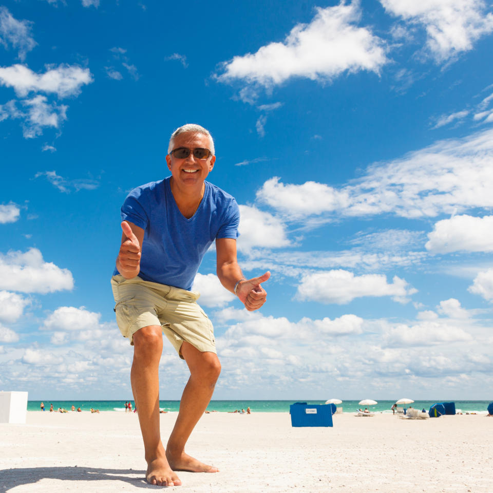 A man in casual beach clothing gives a double thumbs-up while standing on a sandy beach on a sunny day. Blue sky with clouds and ocean in the background