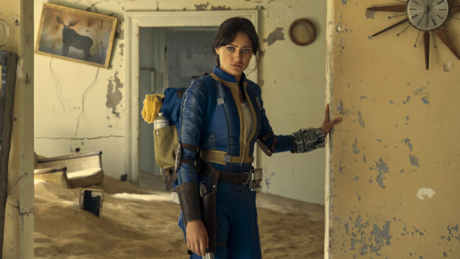 Ella Purnell, as vault dweller Lucy, stands in the ruins of a house where paint is peeling from the walls and inches of sand cover the floor.