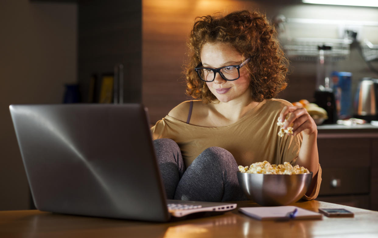 Young woman using computer and snacking on popcorn. (Vesna Andjic / Getty Images)