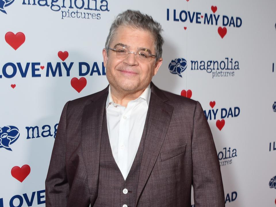 patton oswalt smiling and looking at the camera with his head tilted. he's wearing a brown suit with blazer and vest over a white shirt, as well as frameless glasses