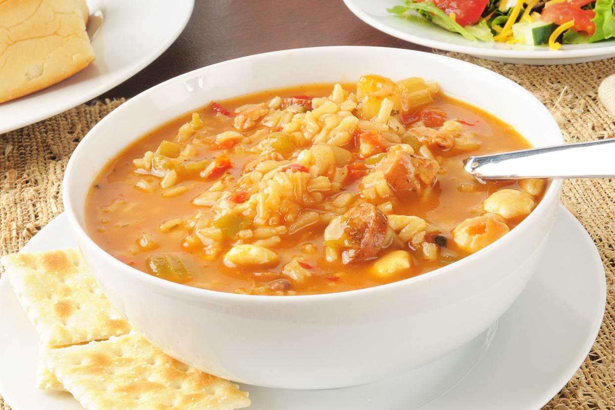 Cajun chicken gumbo in a white bowl with a spoon on a matching white plate with a side of crackers on a napkin with a salad and bread in the background