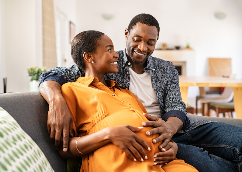 A young Black couple sits on a couch smiling, as the man lays his hand on the pregnant belly of his partner