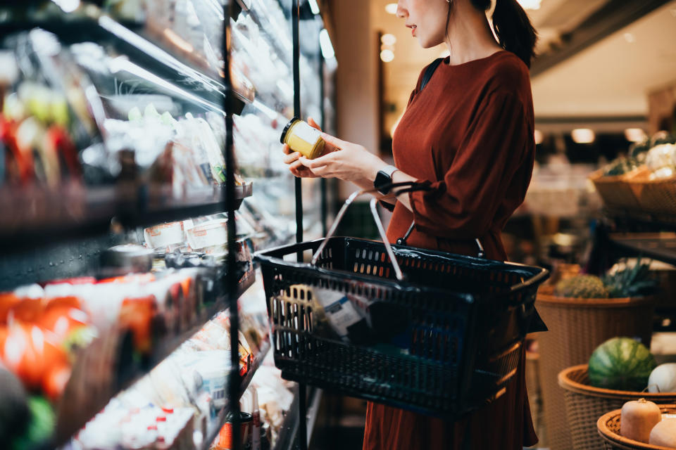 Person shopping, holding a product near shelves with groceries, basket in hand