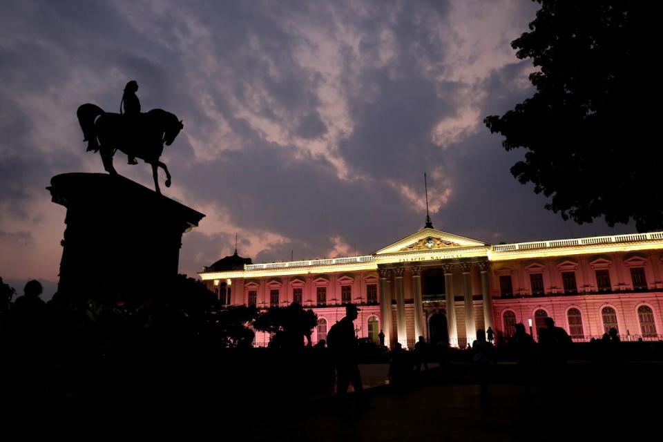 A statue of a man atop a horse, seen in silhouette, in front of a building illuminated in warm orange-pink hues at night