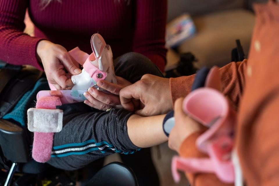 Jewel Calleja, of Livonia, and her husband, Phil Calleja, put ankle braces on their daughter, CC Calleja, 4, as she is secured in a stander to help with her leg strength, circulation and digestion in the living room of their home on Dec. 22, 2023. CC was born with Trisomy 18. Many physicians advise families that children born with Trisomy 18 won't make it out of the hospital, but CC is 4 and her family is finding ways to meet her medical needs.