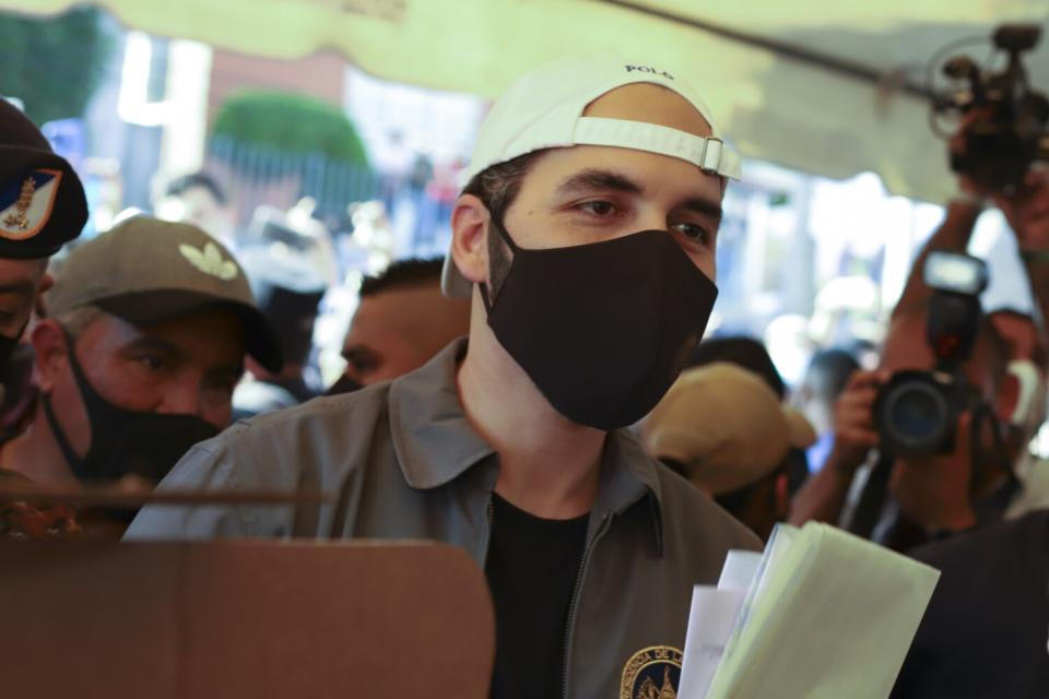 A man in a black mask and white cap worn backward is surrounded by other people.