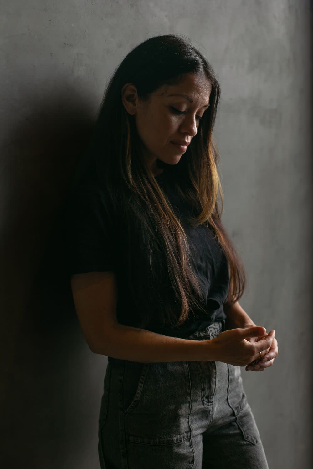 A woman standing with her back against a gray wall, looking down at her hands