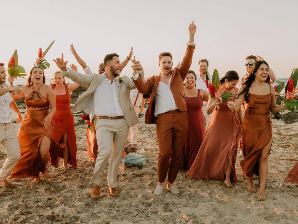 Two grooms raise their hands in celebration with their wedding party on a beach.