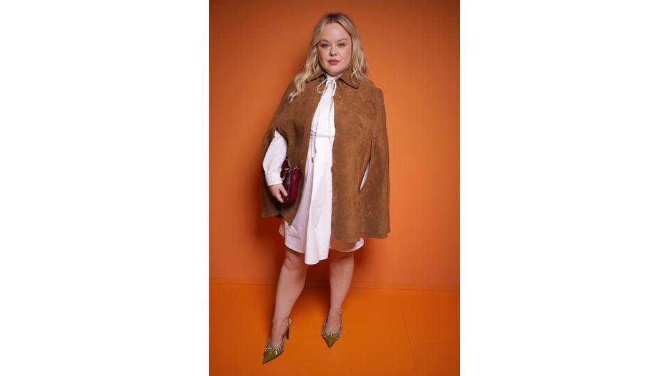 Nicola Coughlan wears an elegant, oversized brown suede cape over a white mini-dress, adding a sophisticated touch to her look. She holds a burgundy handbag in one hand, and her outfit is completed with pointed green heels adorned with crystal embellishments. She exudes confidence with soft makeup and wavy blonde hair.