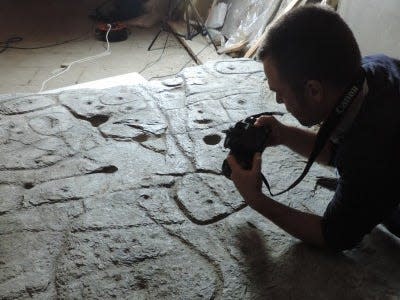 An engraved slab is shown placed on the floor, a scientist leans over it holding a camera. The slab is bigger than the scientist.