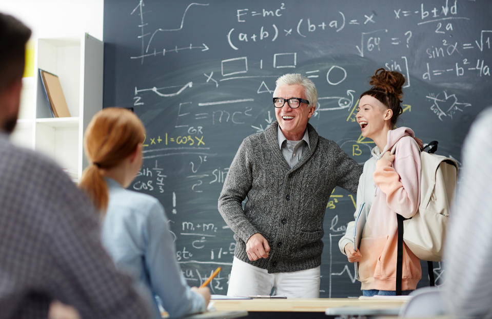 A professor and his student laugh while standing in front of a chalkboard at the front of the classroom