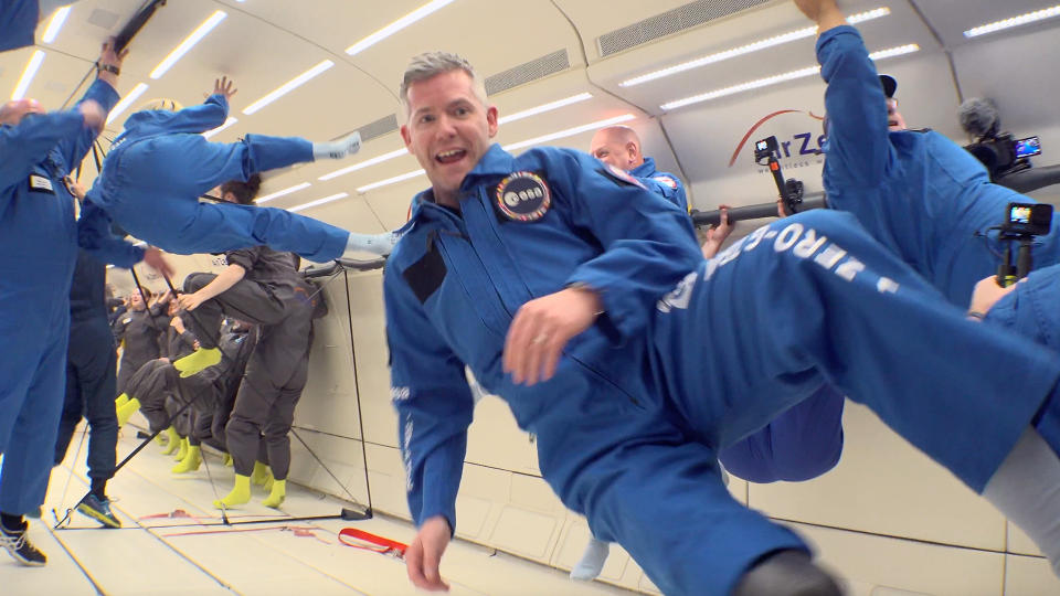     An astronaut in a flight suit floats in front of the camera during a parabolic flight. Behind him, numerous other astronauts in flight suits turn in different directions. 