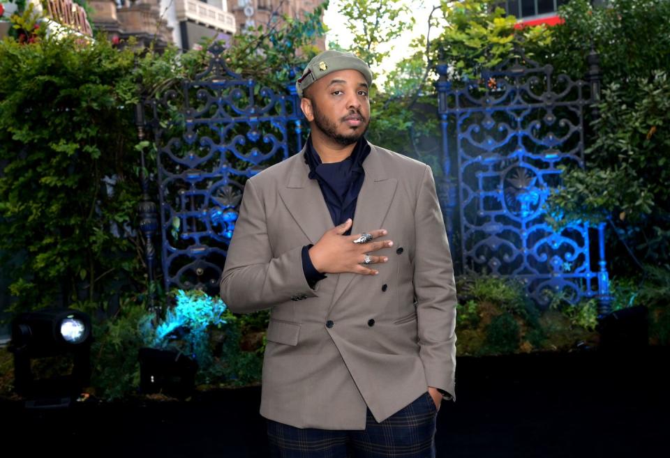 justin simien attends a special screening of haunted mansion, justin is wearing a grey jacket and beanie hat and looking at the camera, with a set of blue gates behind him