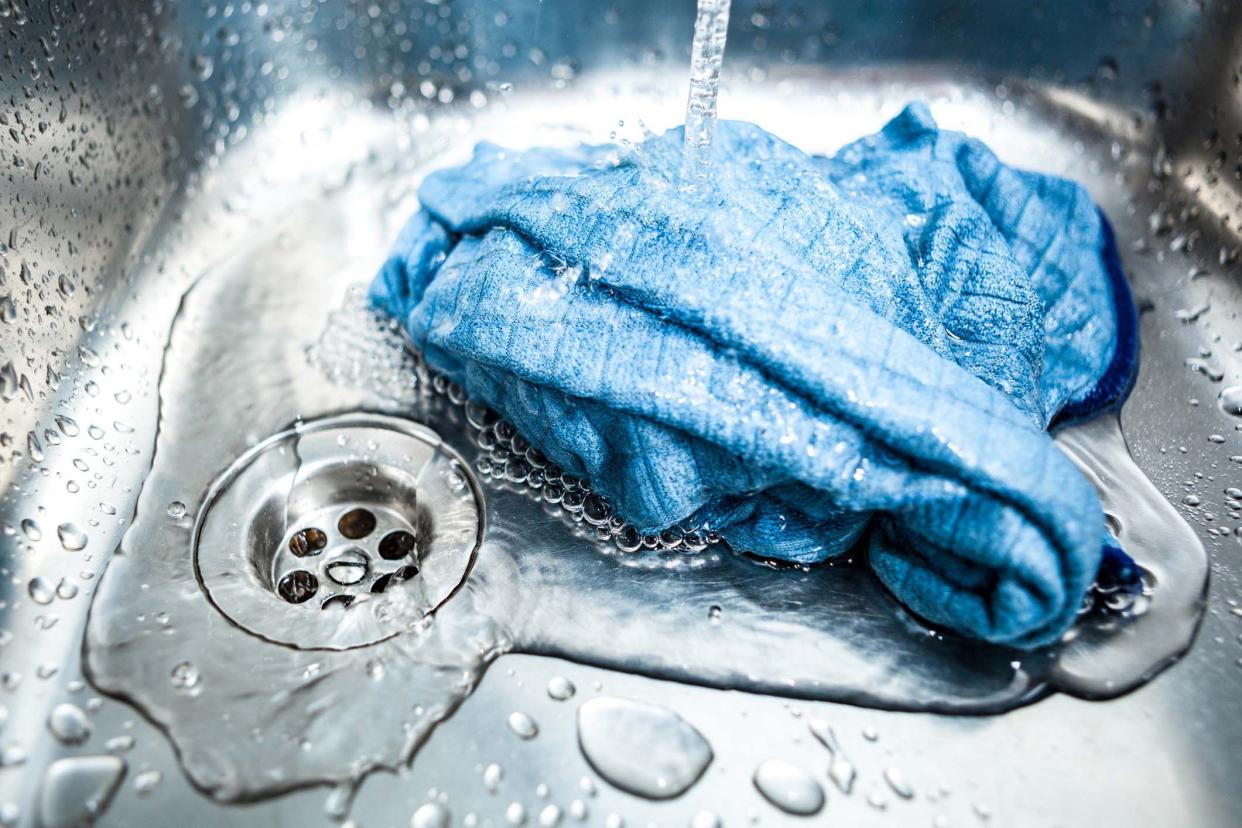 Focus on blue microfiber cloth in a pile under running water in a kitchen sink, drain to the left