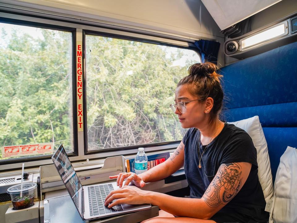 The author uses her laptop while sitting in a blue seat with a window showing greenery outside on her right