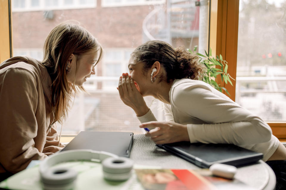 Two women sit at a table, one whispering to the other while smiling. They appear to be sharing a secret.