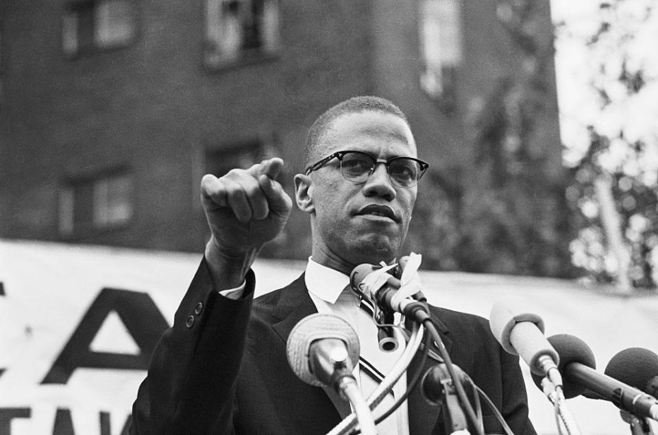 malcolm x speaking into several microphones and points with one finger as he stands outside, he wears glasses, a suit jacket and collared shirt