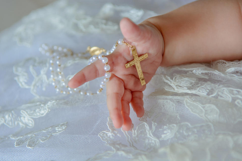 An infant's hand holding a gold cross necklace, resting on delicate lace fabric