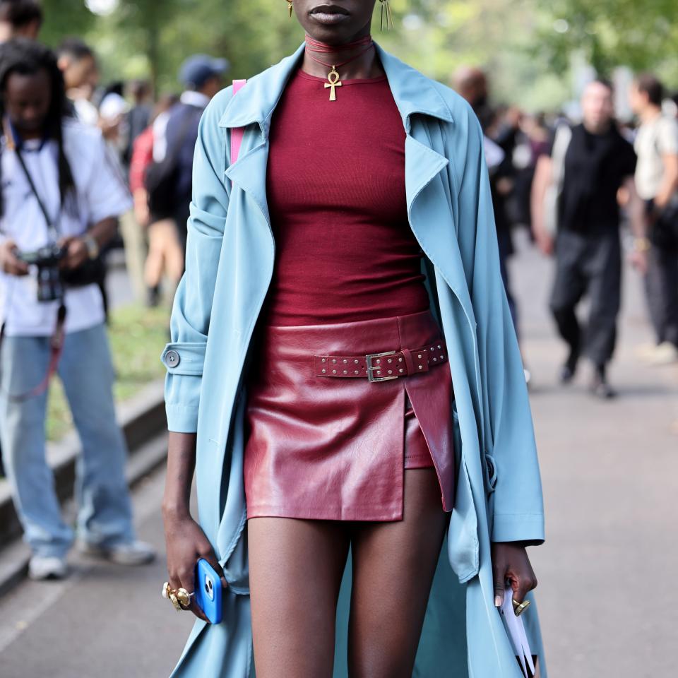 Model wearing a blue trench coat, burgundy top, and leather mini skirt, accessorized with sunglasses and statement jewelry during a street style event