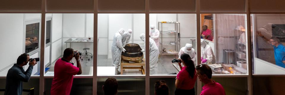 photographers stand behind a glass wall as people in white hazmat suits touch a capsule