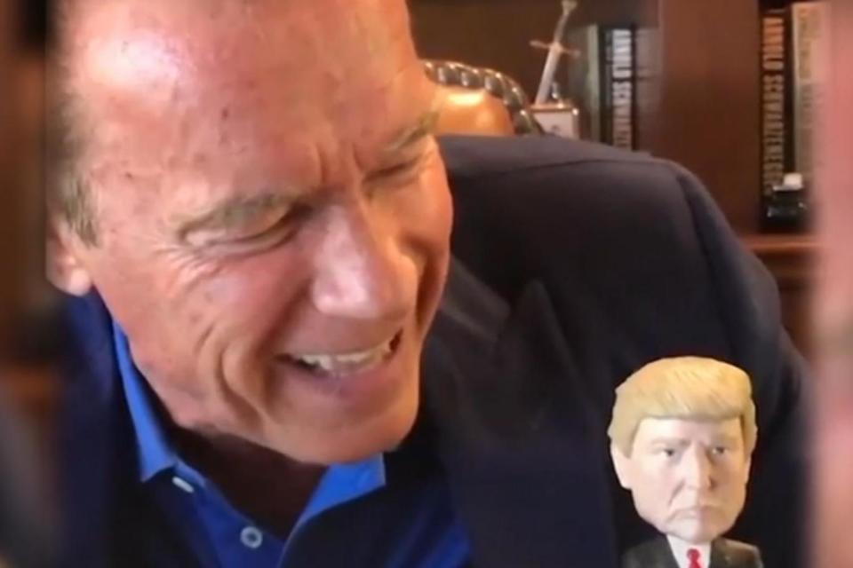 Arnold Schwarzenegger laughs at the nodding Trump toy on his desk