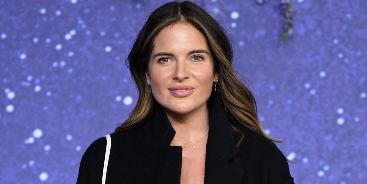 binky felstead, a woman stands looking and smiling at the camera, she wears a black dress and long black coat