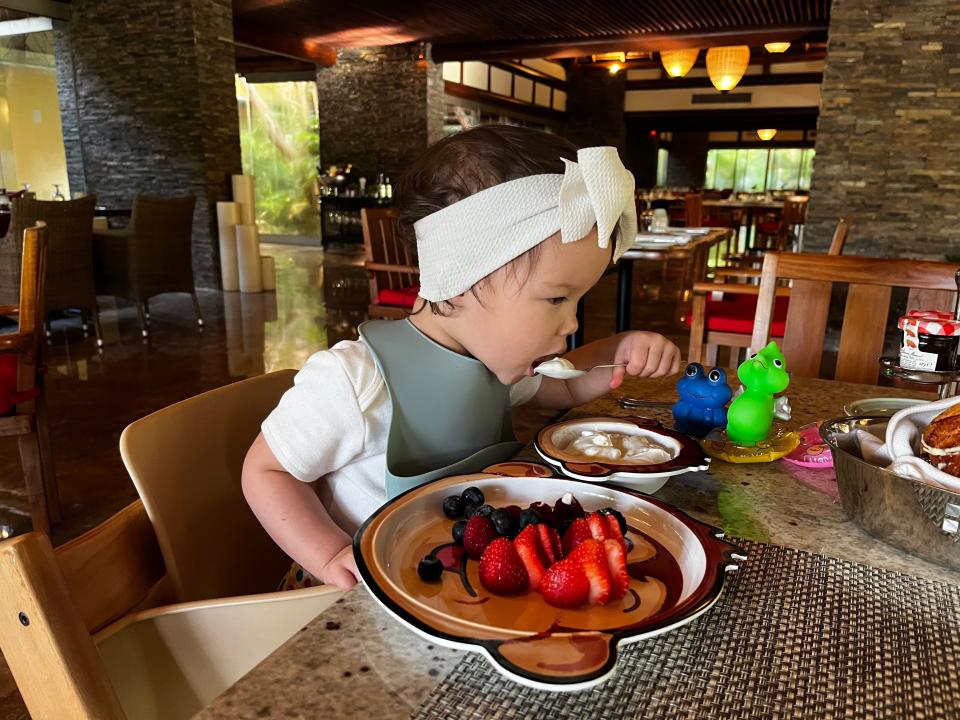 A child wearing a headband a bib eats a spoonful of yogurt with a plate of strawberries and blueberries in front of her