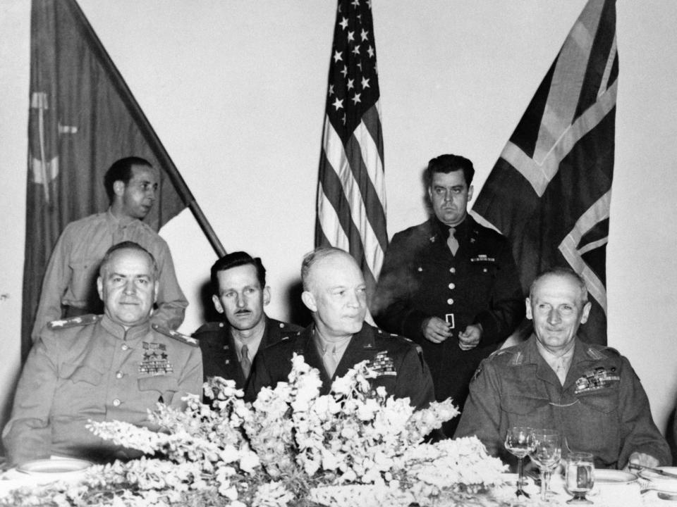 Generals Bernard Montgomery Dwight Eisenhower and Georgy Zhukov sit at a table with flags in the background and a floral arrangement in front