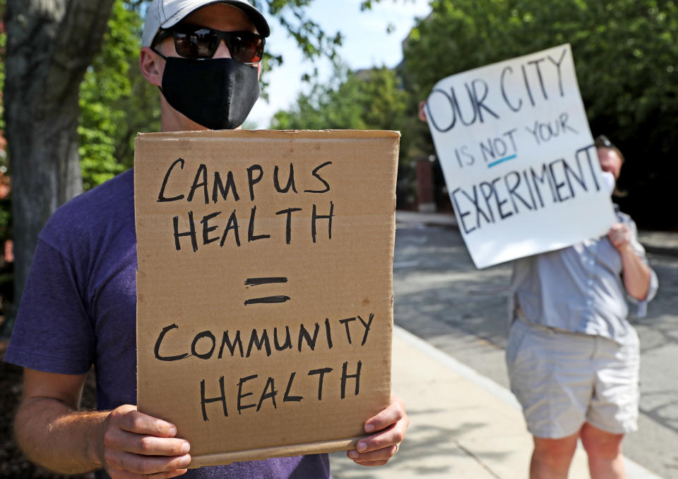 Protesters hold signs reading "Campus health = community health" and "Our city is not your experiment."