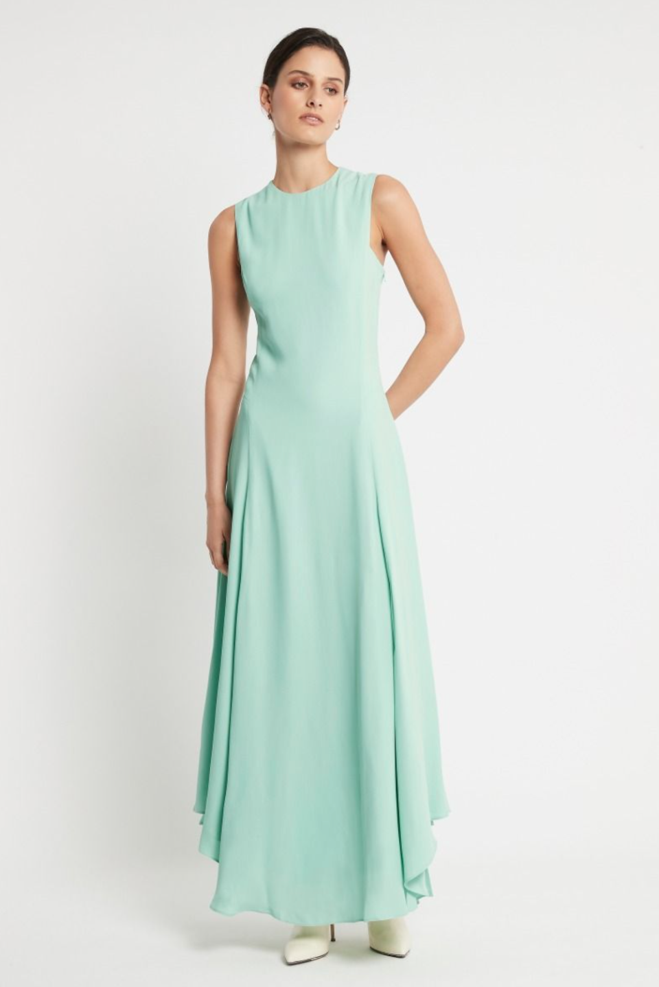 A model wears a long sleeveless gown with flared skirt in mint green, against a white background. She has one hand behind her back and is looking away from the camera.