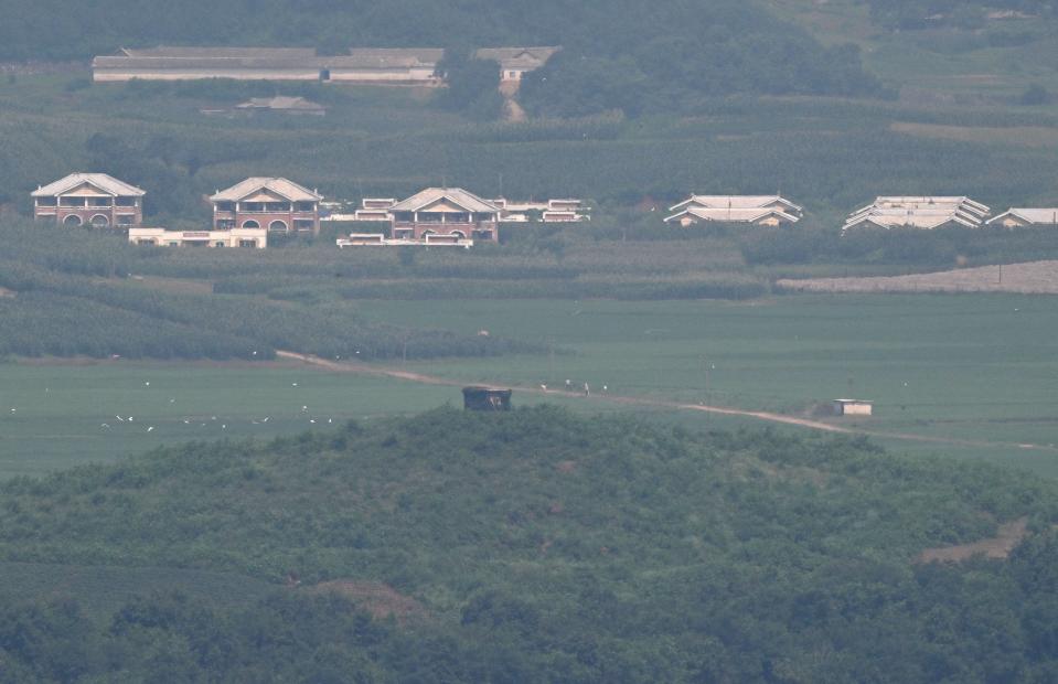 Road crossing through two grassy fields among trees, with several buildings visible in distance.