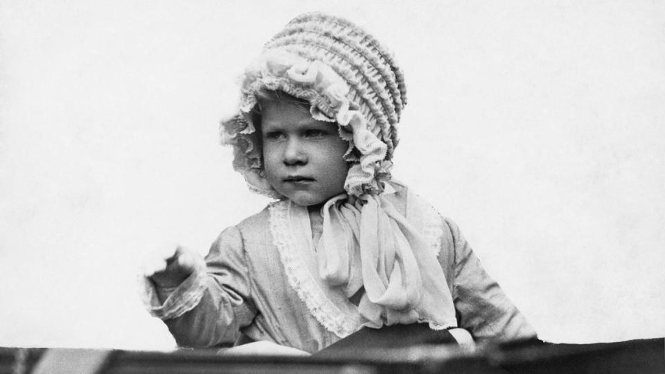 princess elizabeth as a baby sits and waves, she wears a ruffled bonnet and a long sleeve dress